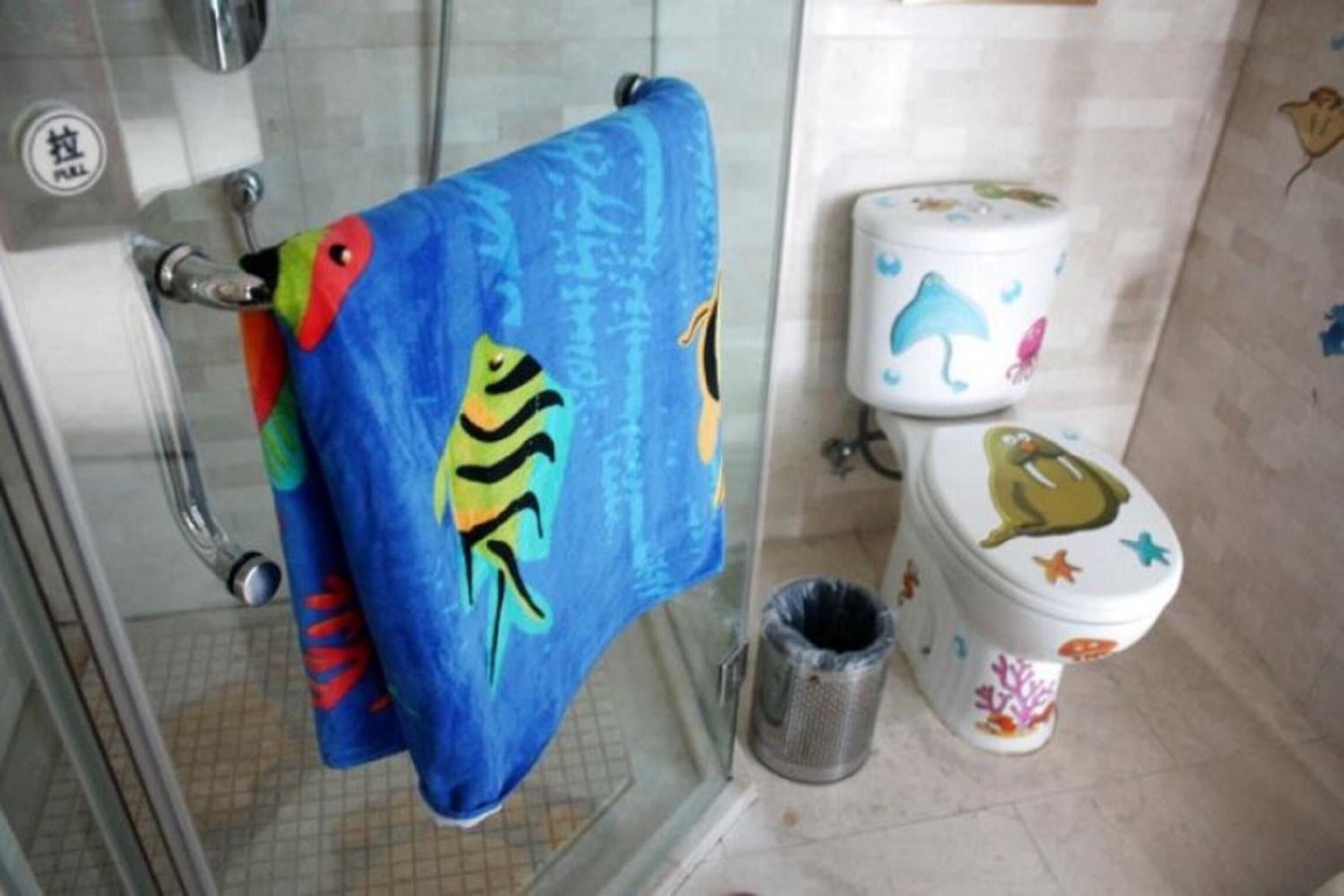 Un baño con ducha, un inodoro decorado con diseños de peces coloridos y un bote de basura.