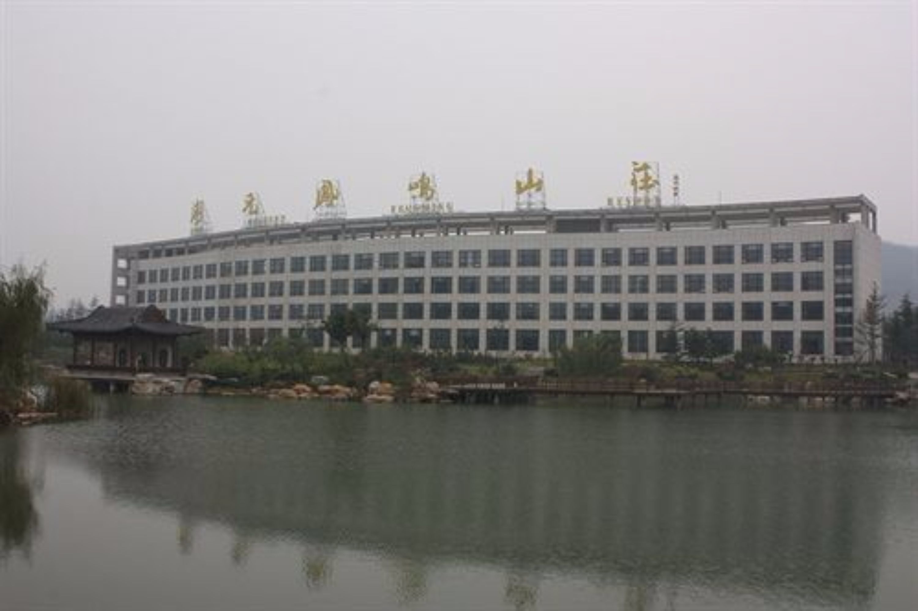 A large, multi-story building with a symmetrical facade, a small pavilion-like structure to the left, and a calm body of water in the foreground.