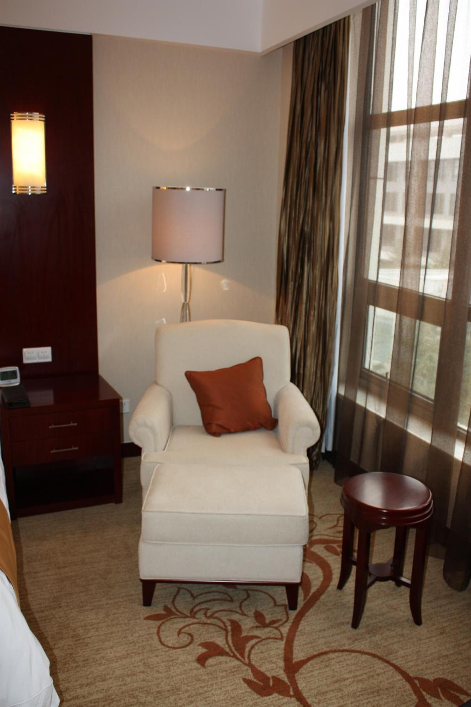 A hotel room with a white armchair, a small wooden side table, a lamp, and a large window with curtains.