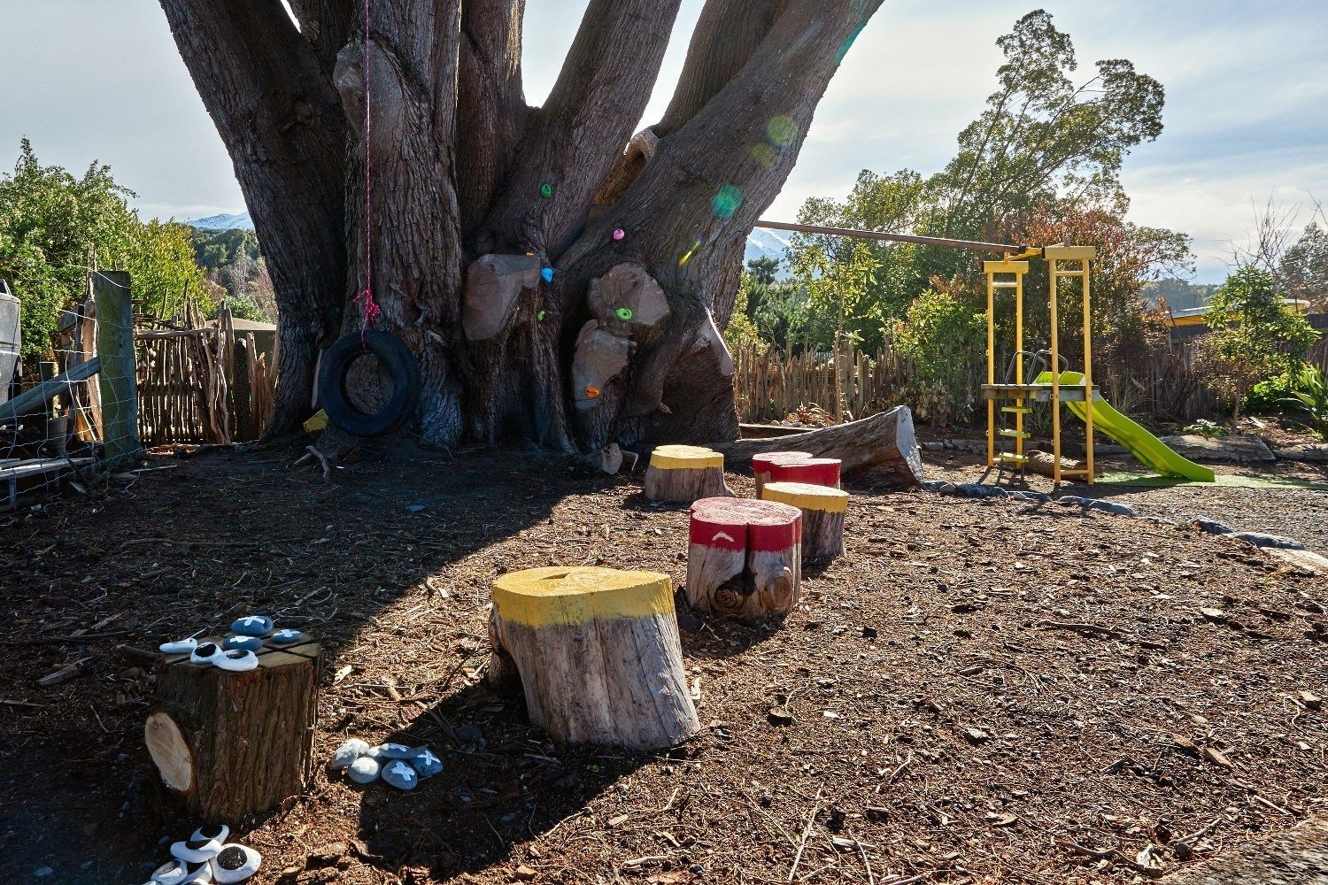 Une aire de jeux avec un grand arbre, des troncs d’arbres et une structure de jeu jaune.
