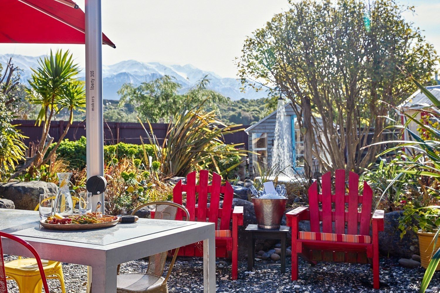 Een terras buiten met een eettafel, rode stoelen, een metalen emmer met ijs en een bergzicht op de achtergrond.