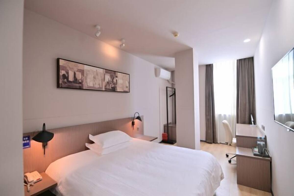 A hotel room with a bed, bedside table, lamp, desk, chair, and a large window.