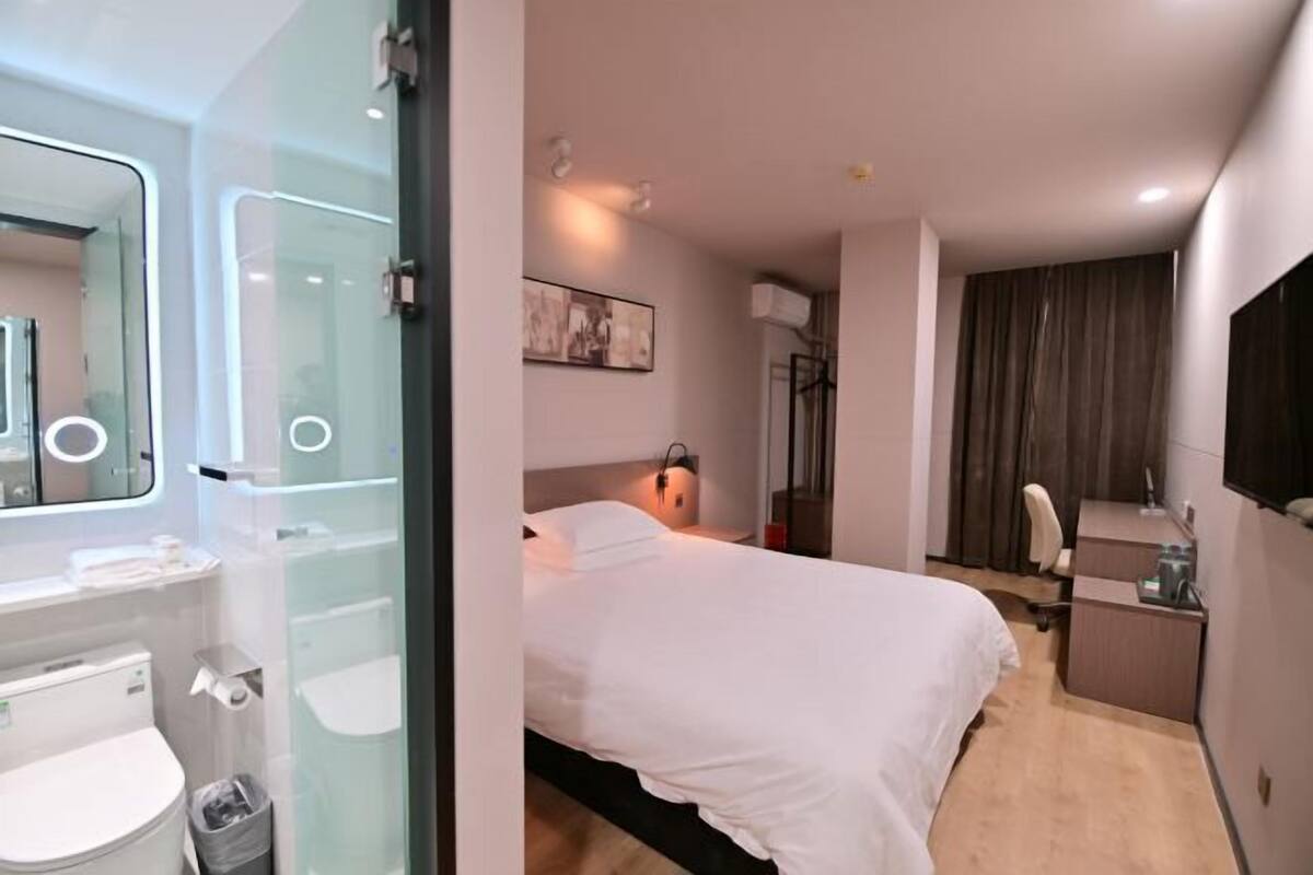 A modern hotel room with a glass shower enclosure, a bed with white bedding, a desk with a chair, and a mounted TV.