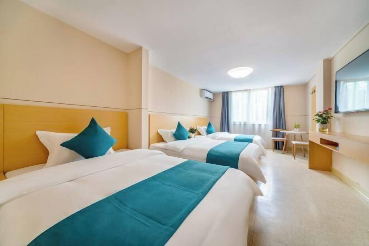 Three beds with white linens and teal accents, a desk area, and a large mirror.