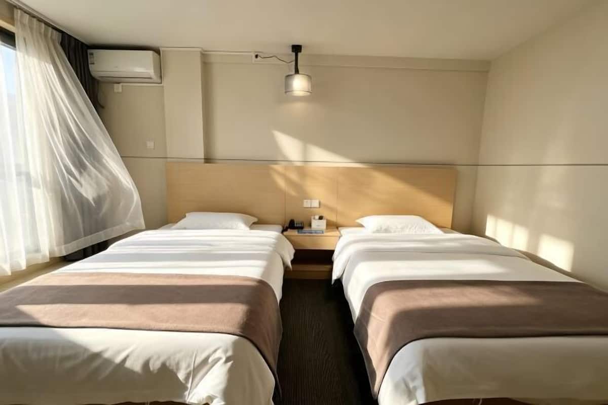 A hotel room with two beds, a nightstand, an air conditioning unit, and a window with curtains.