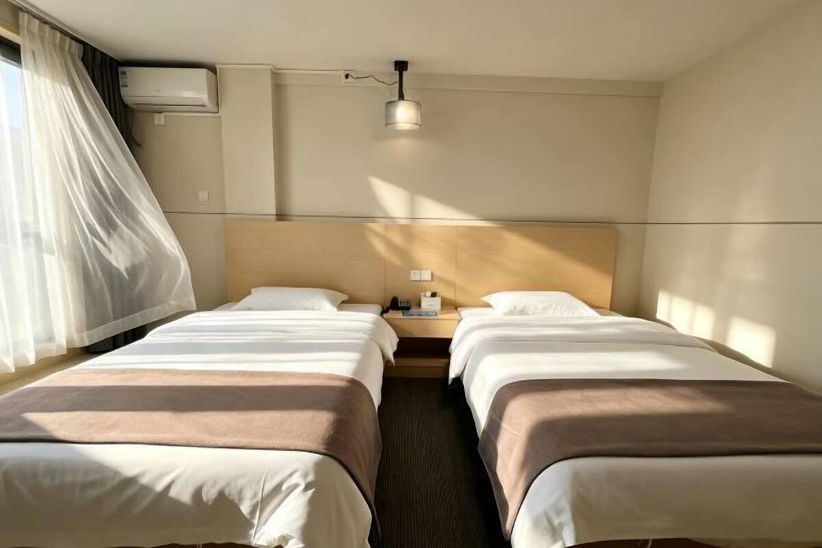 A hotel room with two beds, a nightstand, a lamp, and a window with curtains.
