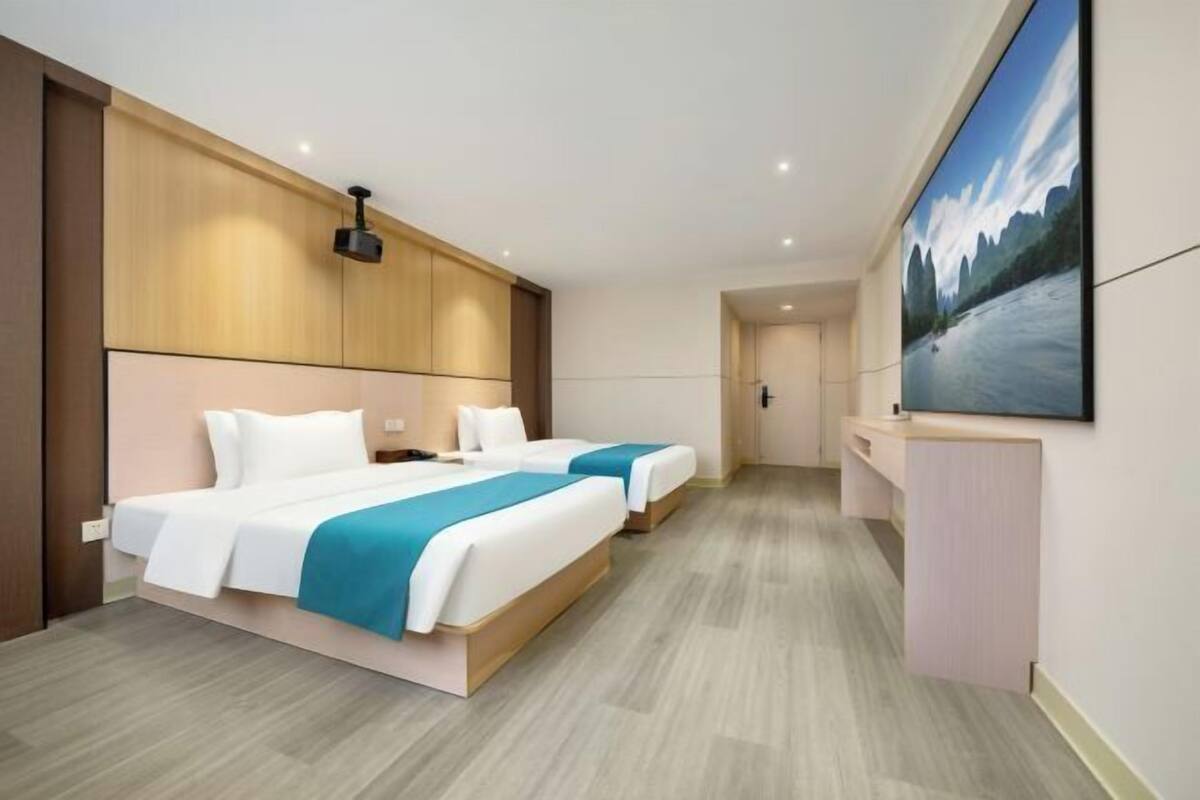 A hotel room with two beds, a desk, a large window, and a painting on the wall.