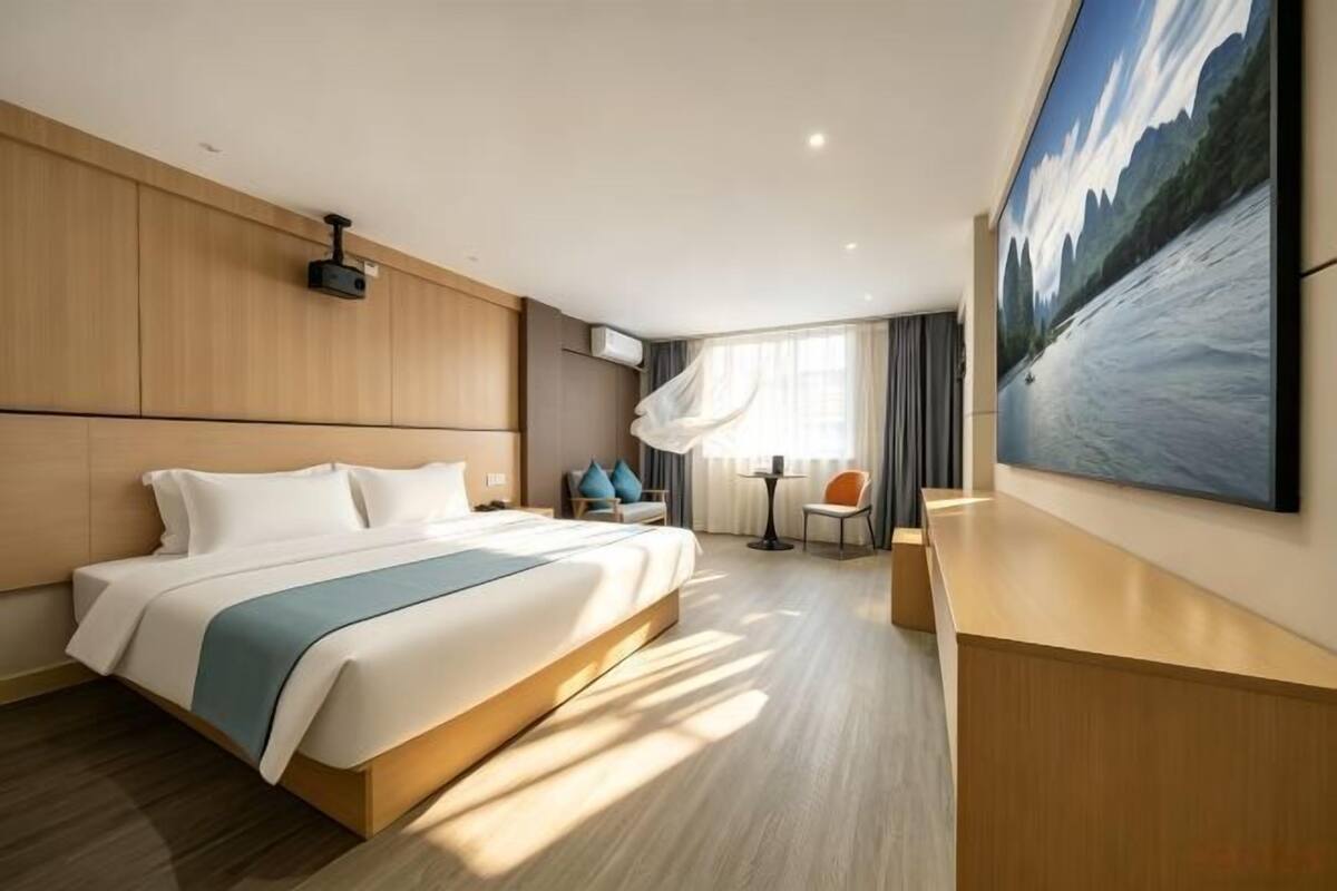 A modern hotel room with a large bed, a desk, a chair, a painting, and a window with curtains.
