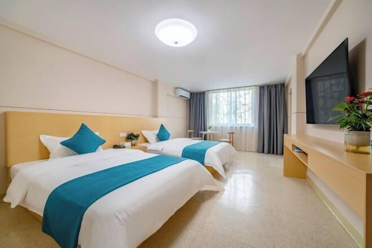 Two beds with white bedding and blue accents, a flat-screen TV, and a small table with a chair near the window.