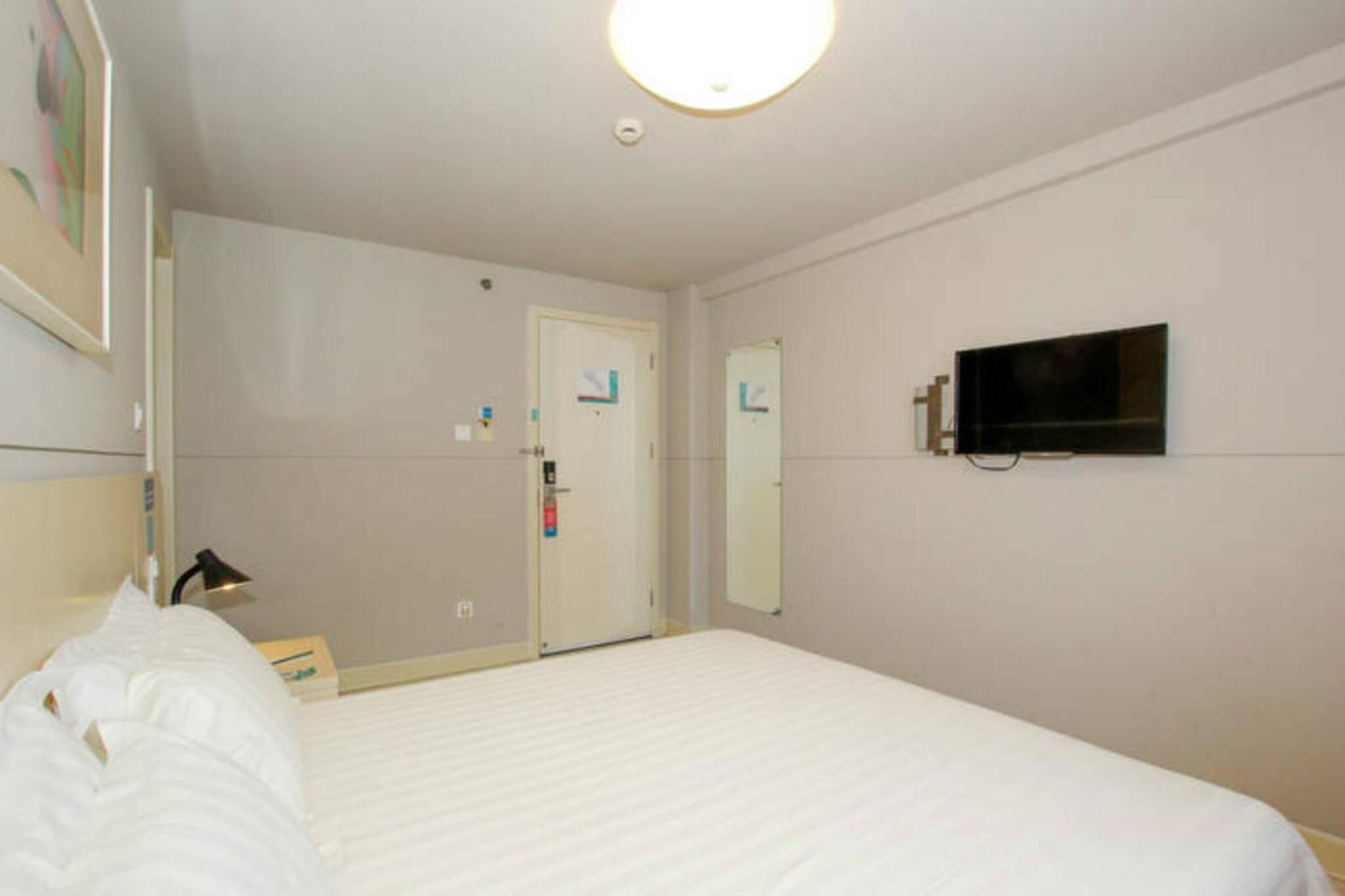 Une chambre d’hôtel avec un lit, un téléviseur fixé au mur, une porte et une petite table avec une lampe.