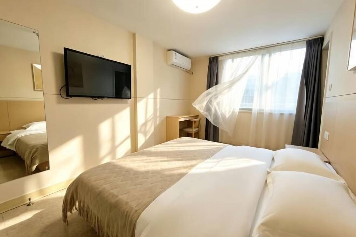 A hotel room with a bed, a TV mounted on the wall, an air conditioning unit, and a window with curtains.
