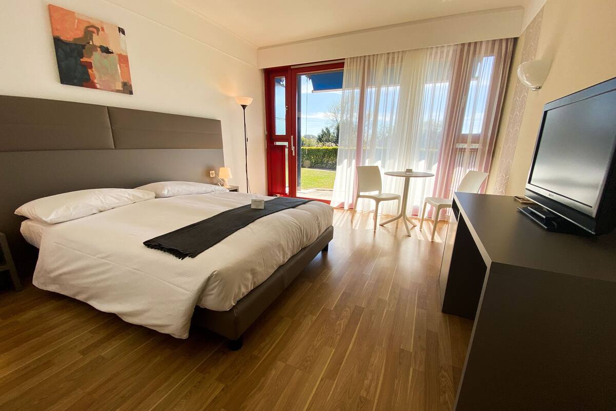 Een hotelkamer met een groot bed, een bureau met een televisie, een kleine tafel met stoelen en uitzicht op het omliggende landschap door het raam.