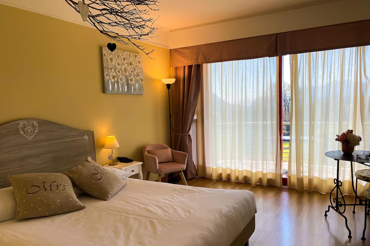 Een hotelkamer met een groot bed, een nachtkastje met een lamp, een stoel, een tafel met een vaas, en uitzicht op het buitengebied door dunne gordijnen.