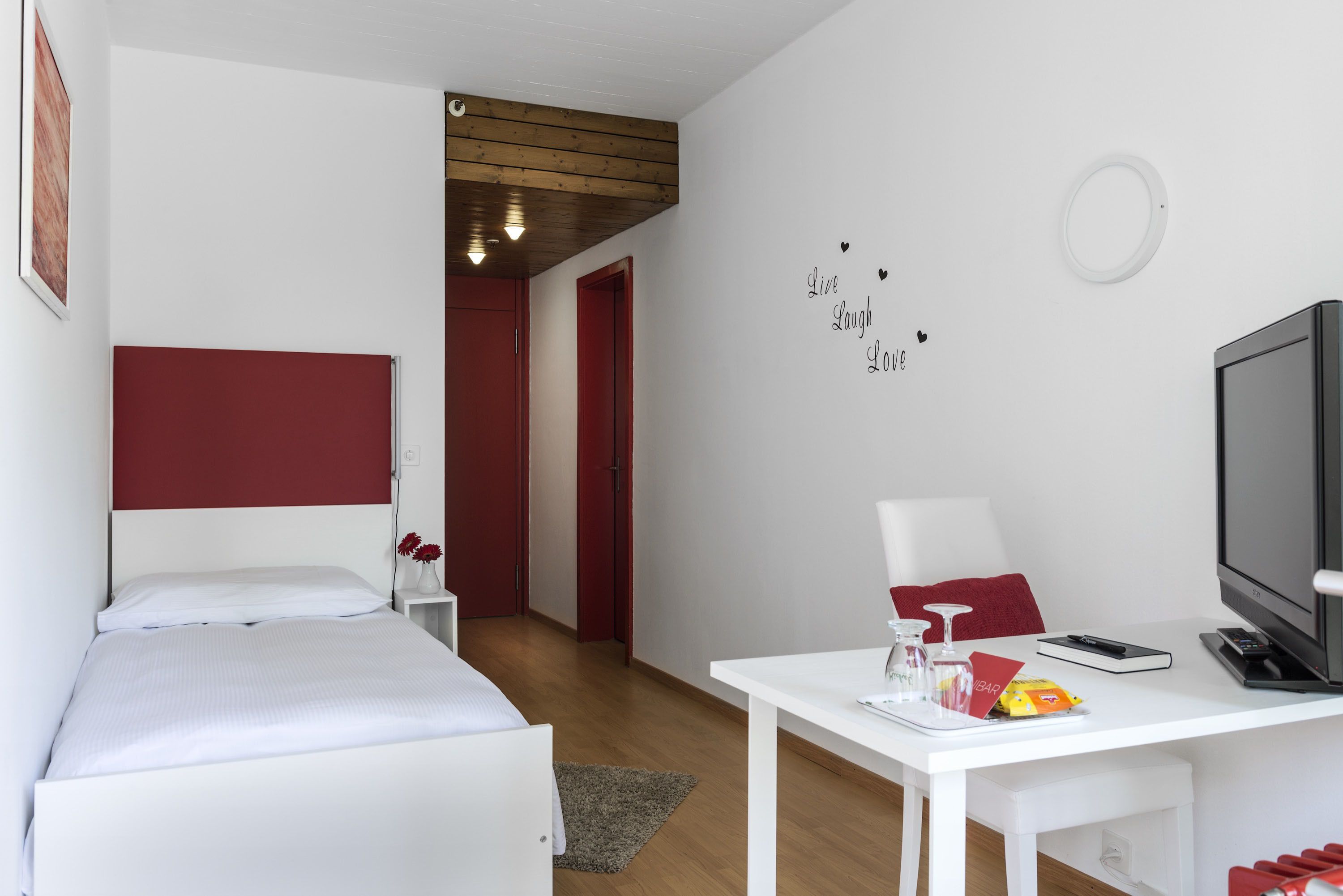 Une chambre d’hôtel avec un lit, un bureau avec une télévision, une petite table avec un verre et un objet rouge, et un mur orné d’une citation inspirante.
