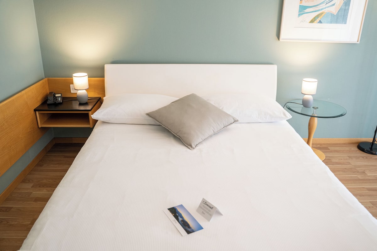 Een net opgemaakt bed met een kussen, een nachtkastje met een lamp, en een klein rond tafeltje met een lamp.