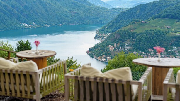 Une terrasse meublée de mobilier en bois, donnant sur un lac et un village.