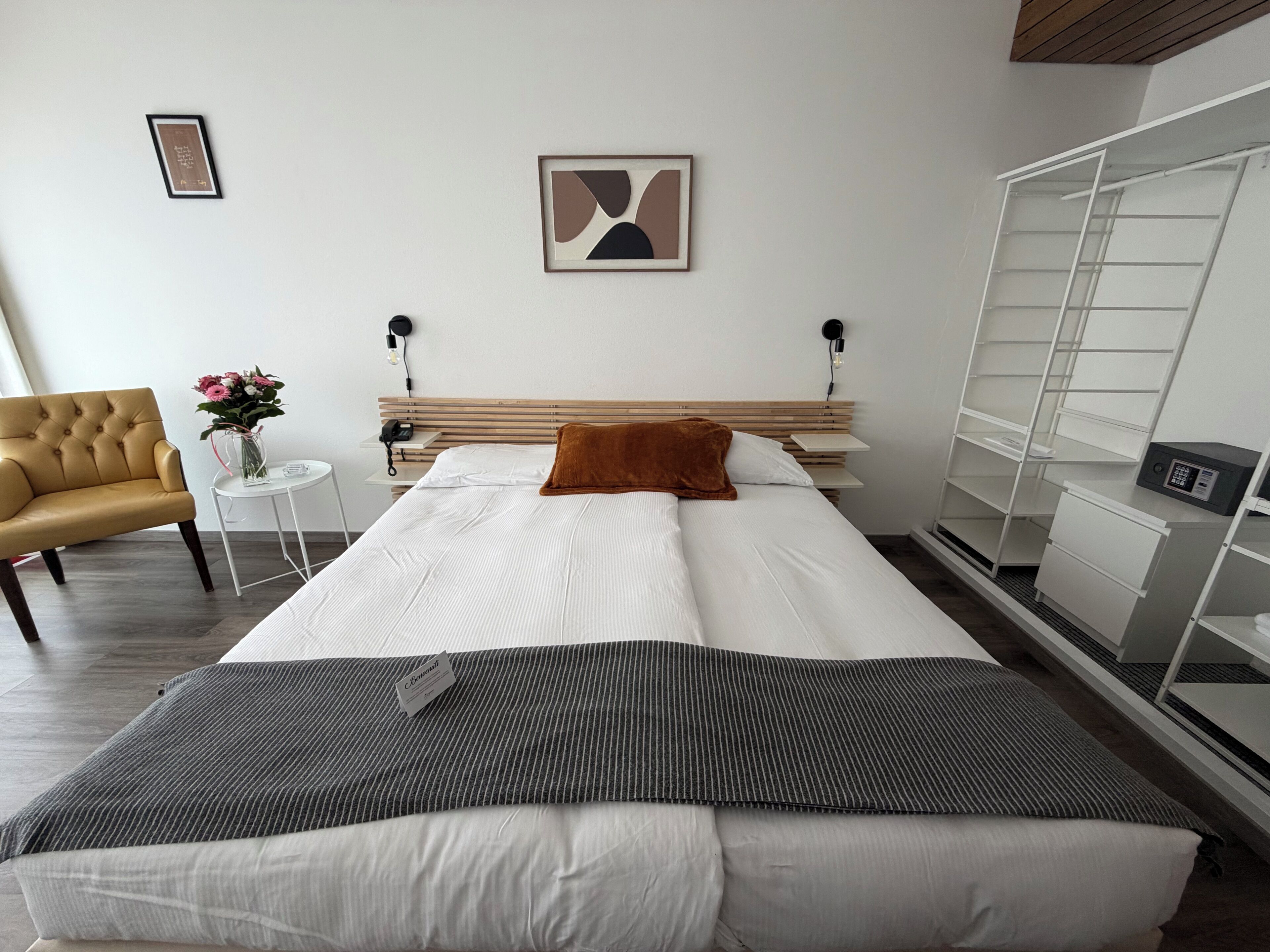 Una camera d'albergo con un letto, una poltrona, un tavolino con un vaso di fiori, un comodino con una lampada, un'opera d'arte appesa al muro e un armadio a muro.