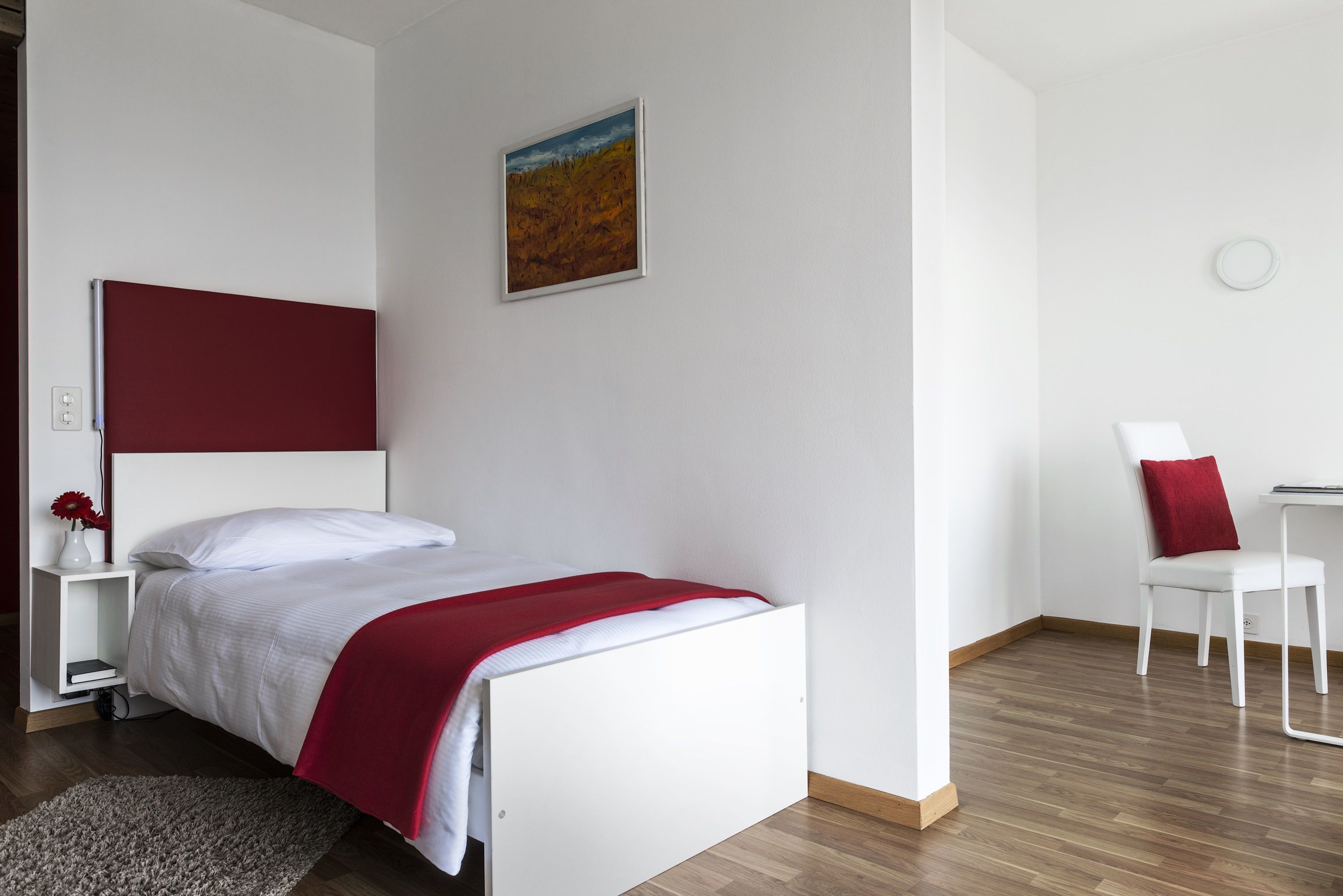 A modern bedroom with a white bed, red accents, a painting on the wall, and a small desk with a chair.
