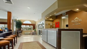 A hotel reception area with a reception desk, bar stools, a large potted plant, and a view of the outside through a glass door. - Microtel Inn & Suites by Wyndham Dickinson (Dickinson)