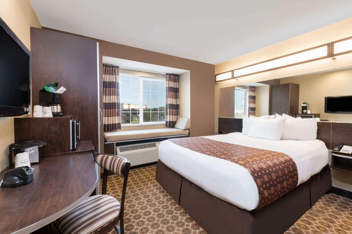 A hotel room with a large bed, a desk with a chair, a television, a window with curtains, and a mirror.