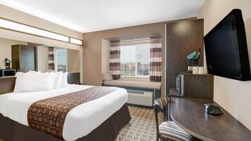 Habitación de hotel con cama, escritorio, silla, televisor, ventana con cortinas y vistas a la ciudad.