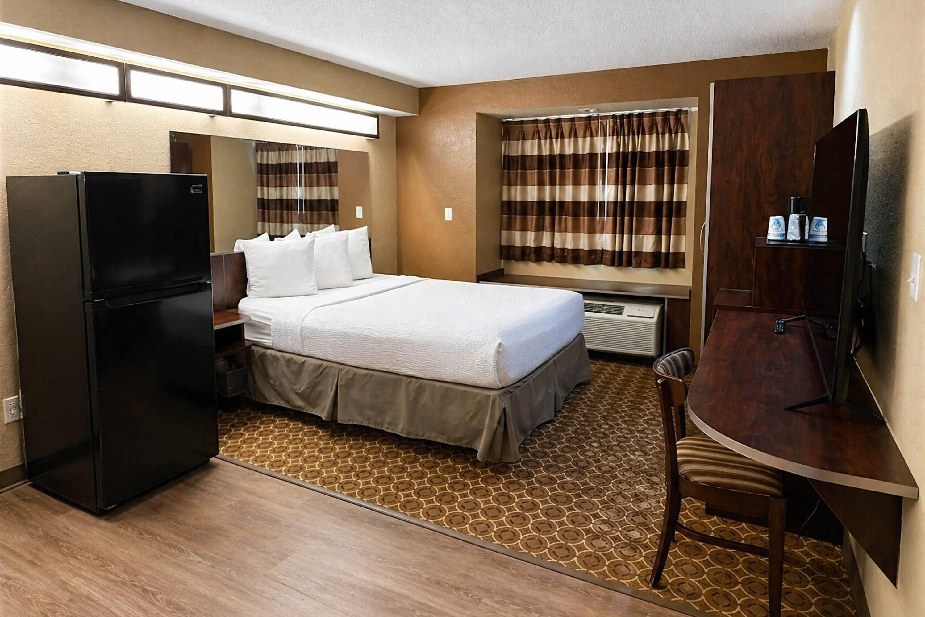 A hotel room with a bed, a refrigerator, a desk, a chair, a window with curtains, and a television.