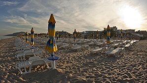 Una playa con filas de tumbonas y sombrillas, una orilla arenosa y edificios al fondo.