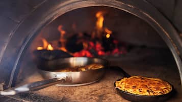 Un horno de pizza con una pizza en su interior, una sartén con comida y llamas al fondo.
