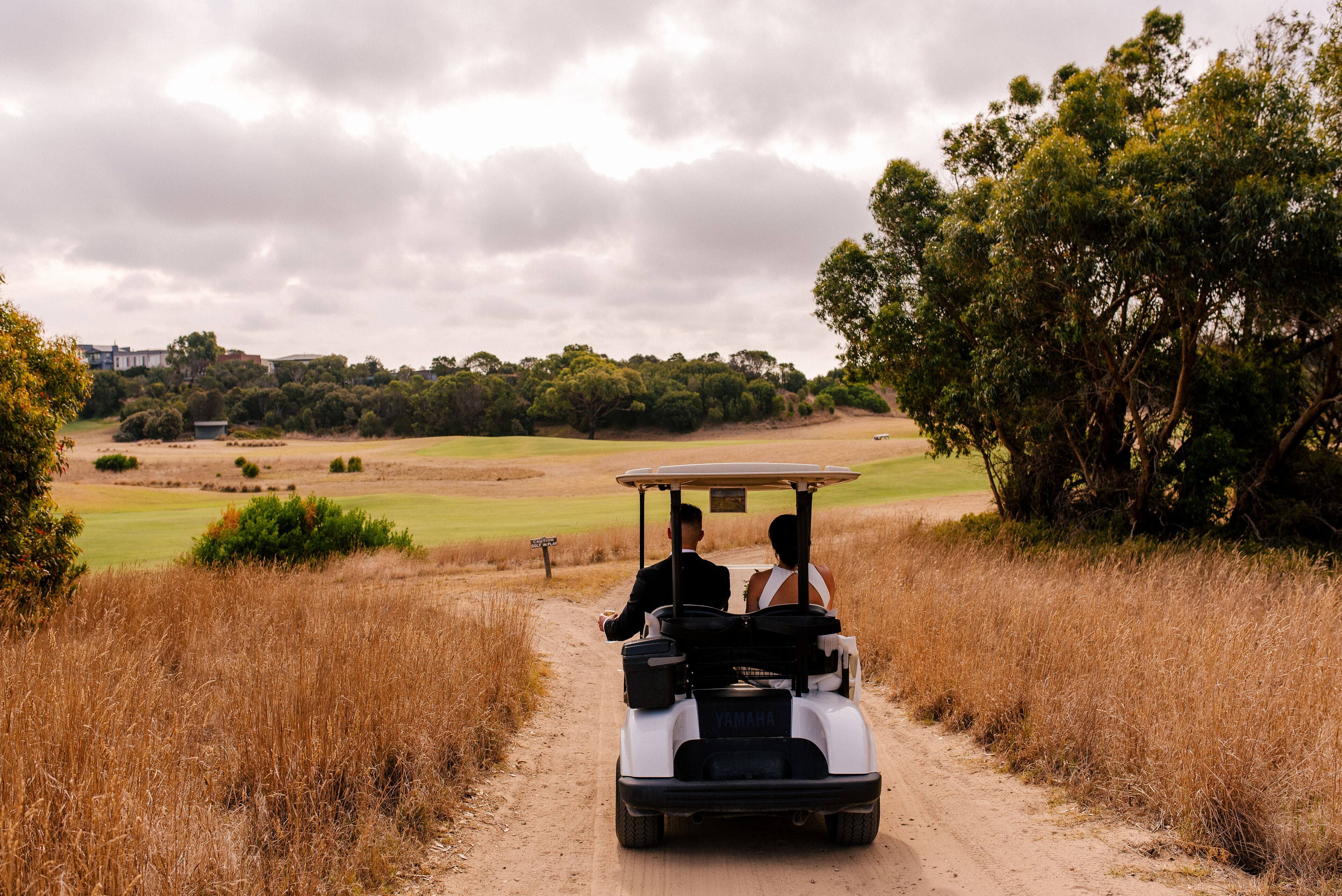 Un carrito de golf en un camino de tierra, con una pareja sentada, rodeado de pasto seco y árboles, y un campo de golf al fondo.