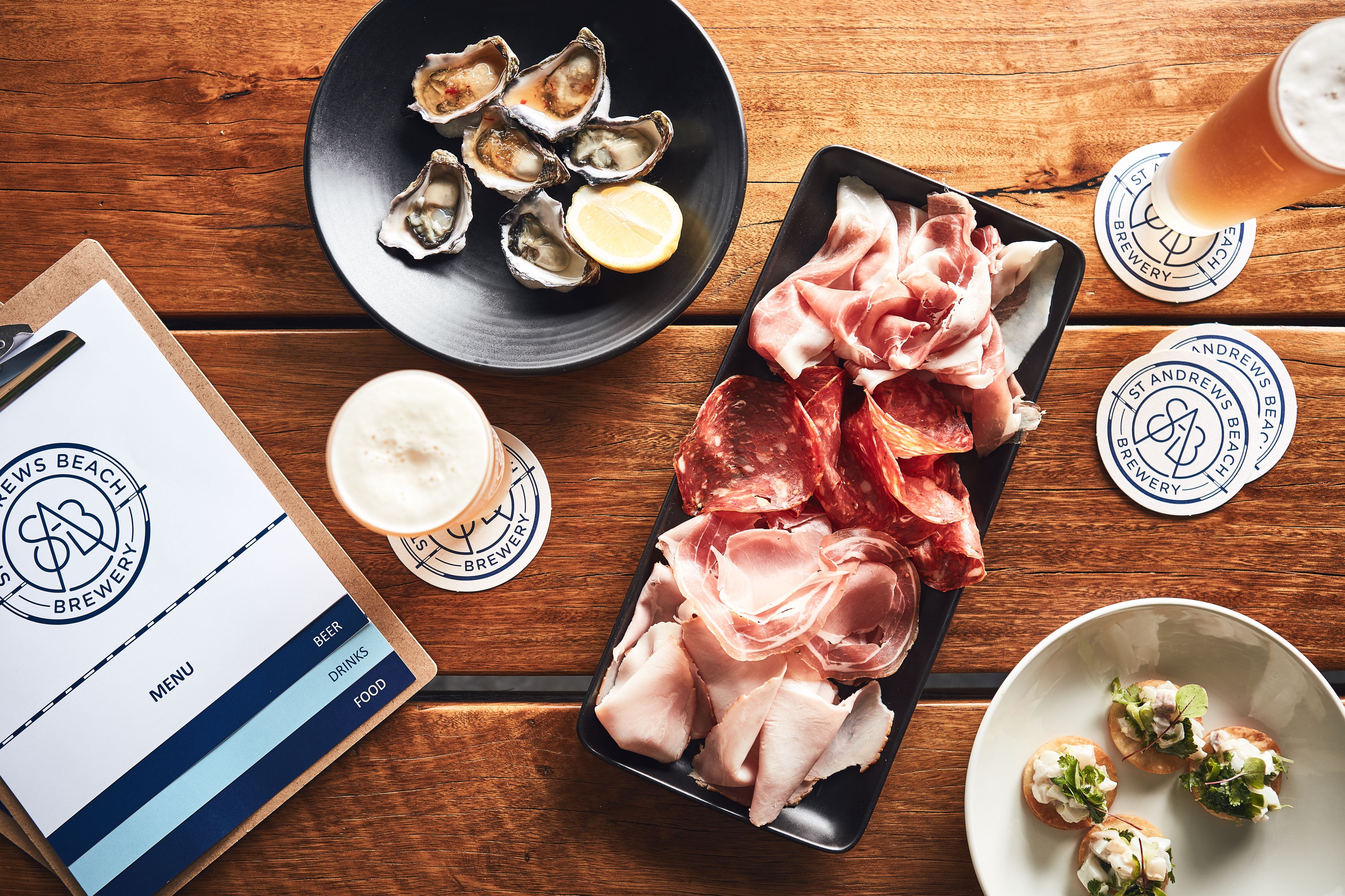 A wooden table with a menu, a plate of oysters, sliced meats, and a glass of beer.