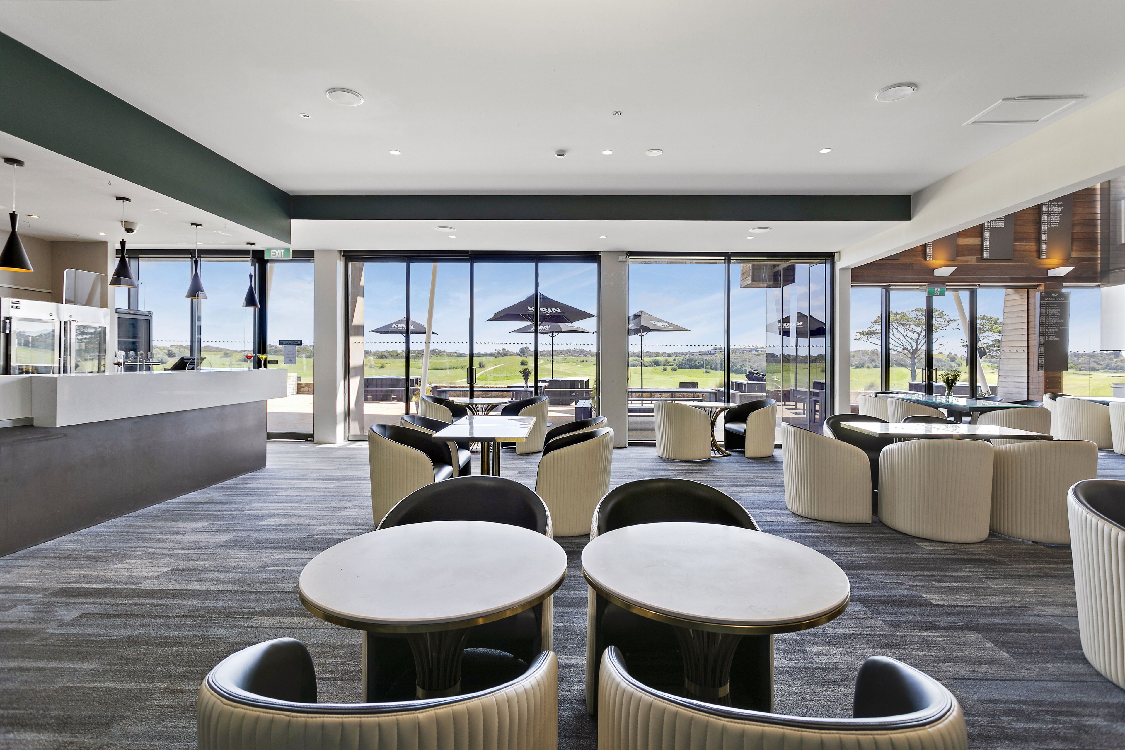 A modern indoor dining area with round tables and chairs, large windows offering a view of the outdoors, and a kitchen area visible in the background.