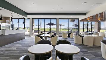 Un comedor interior moderno con mesas y sillas redondas, ventanas amplias con vistas al exterior y una zona de cocina visible al fondo.