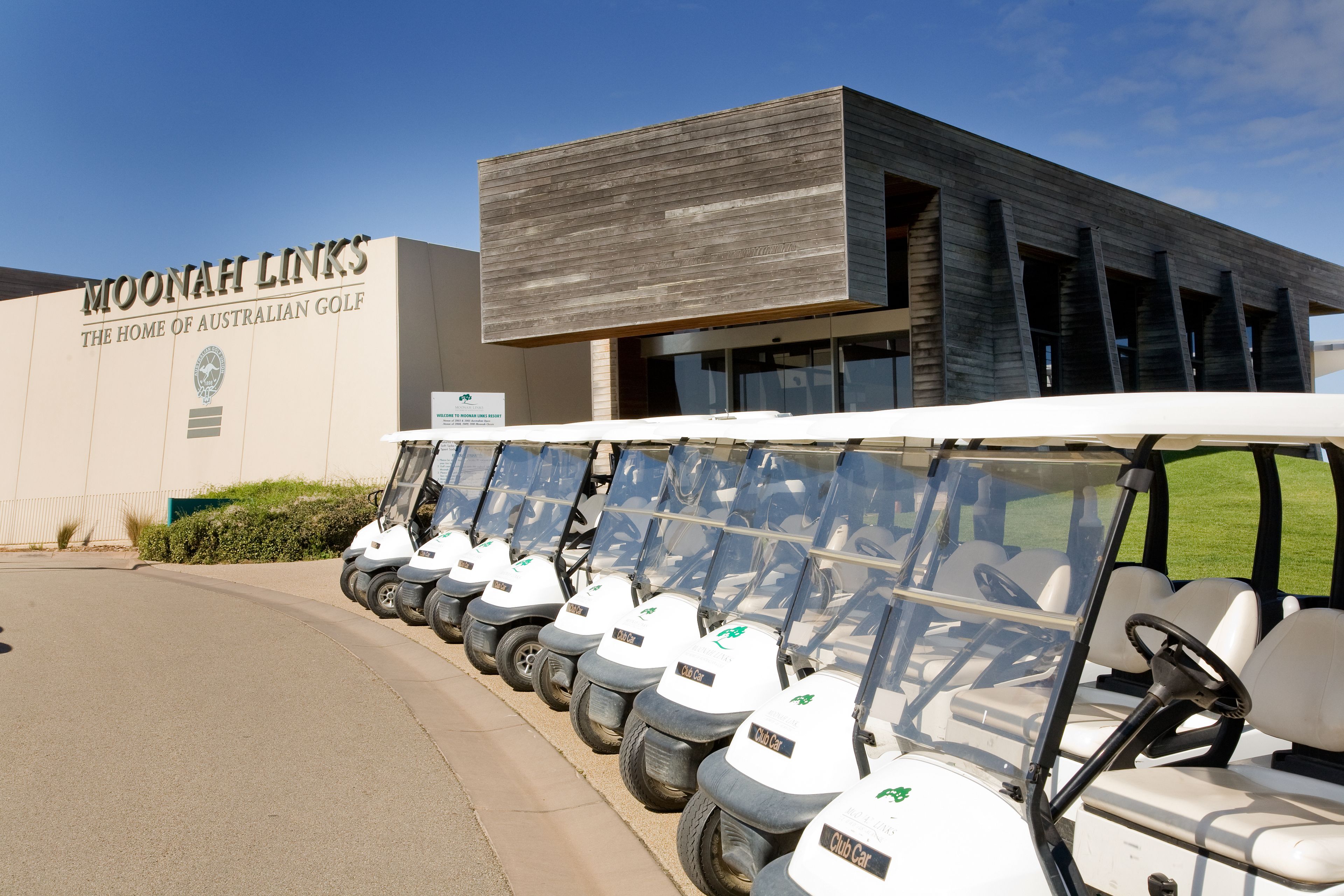 Un edificio con un cartel que dice "MOONAHI LINKS - THE HOME OF AUSTRALIAN GOLF" y una fila de carros de golf estacionados frente a él.