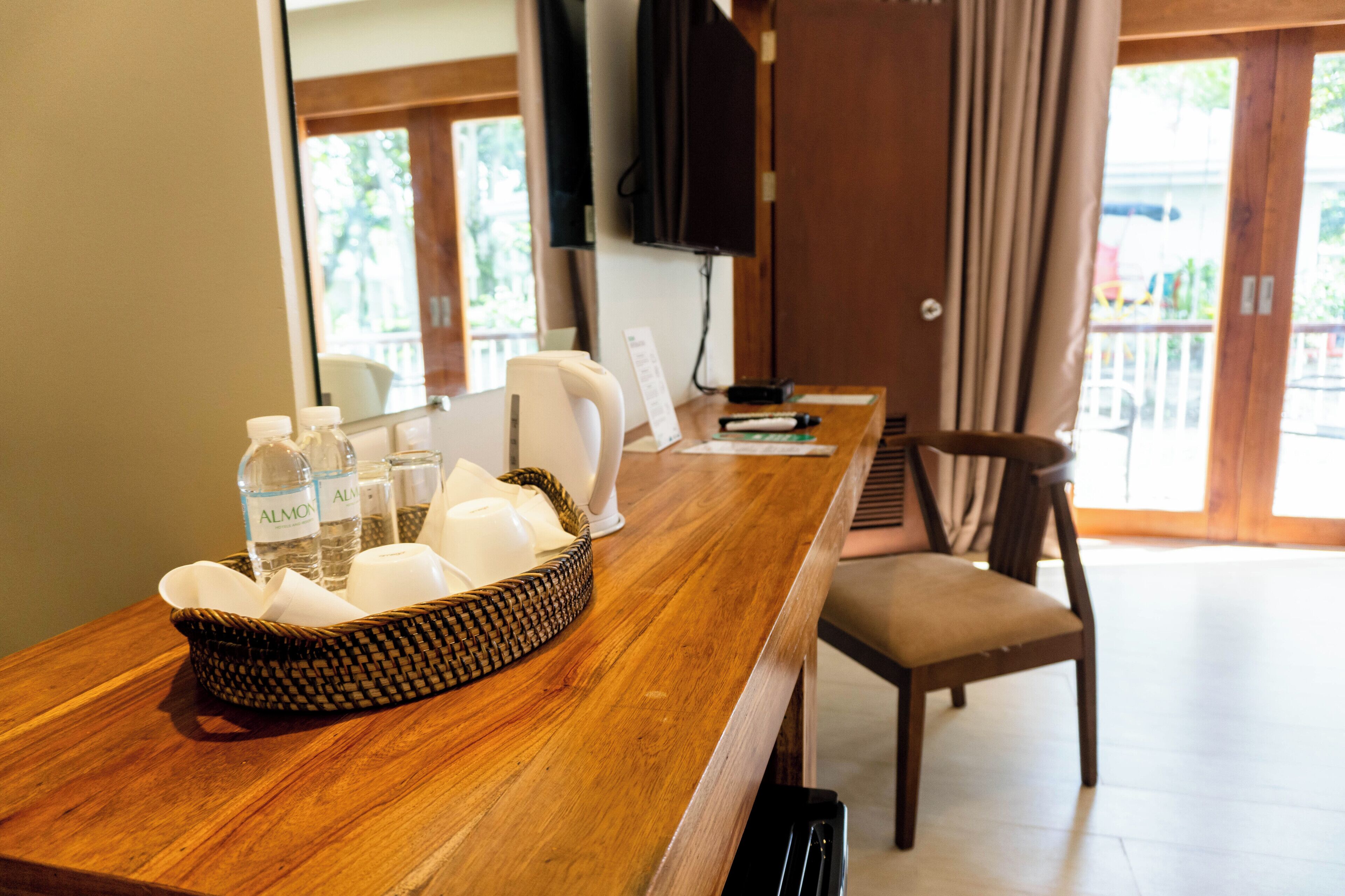 Una mesa de comedor de madera con una cesta tejida que contiene botellas y vasos, una tetera eléctrica blanca y un control remoto. Una silla está colocada al final de la mesa.
