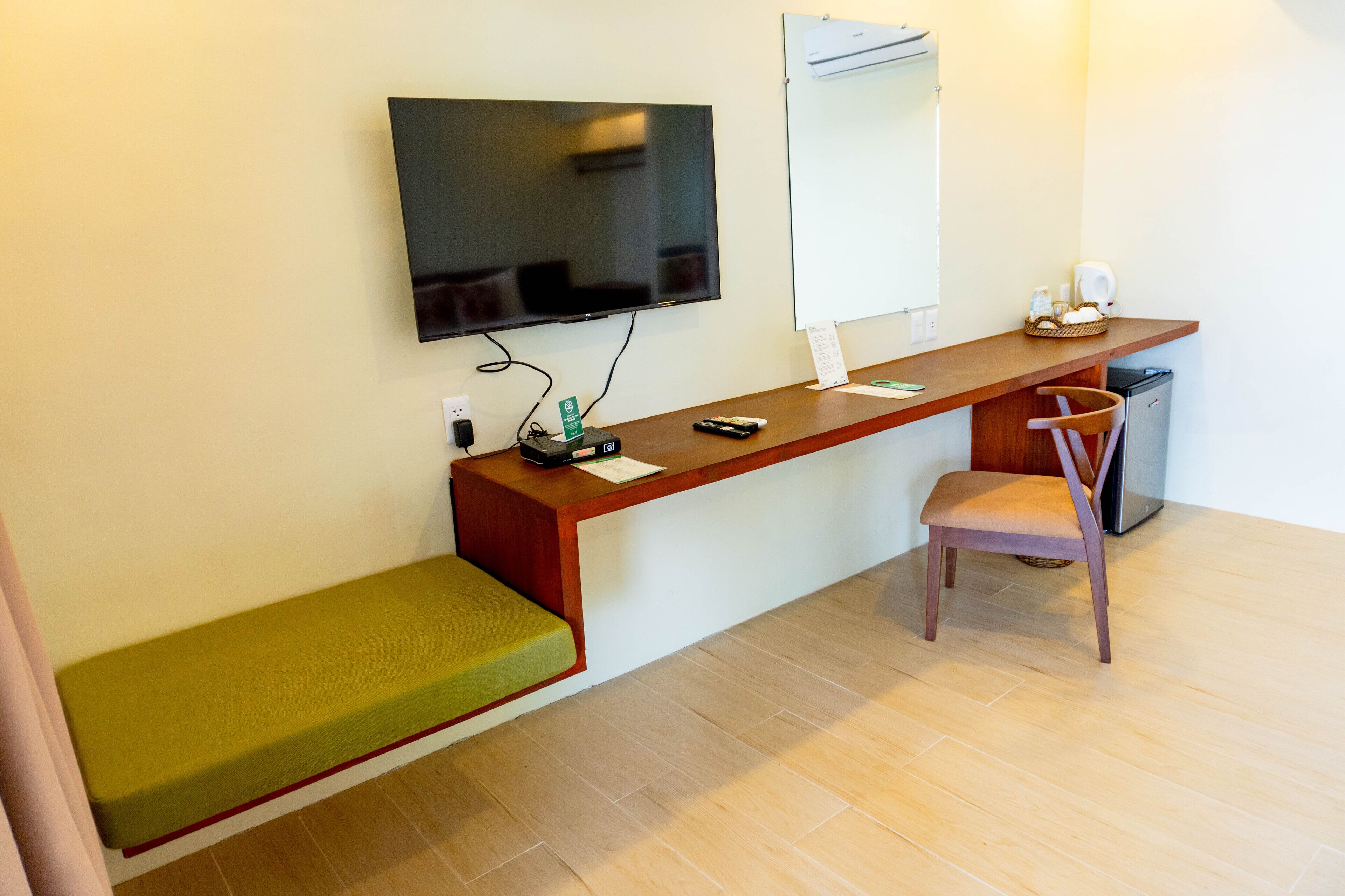 Una habitación con un escritorio de madera, un banco verde, un televisor, una pizarra y un refrigerador.