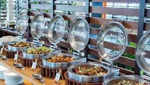 A buffet with various dishes in metal containers, arranged on a wooden counter with a view of greenery through a wooden slatted wall. - Almont Inland Resort (Butuan)