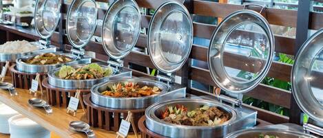 Buffet com diversos pratos em recipientes metálicos, dispostos sobre um balcão de madeira com vista para o verde através de um painel de ripas de madeira.