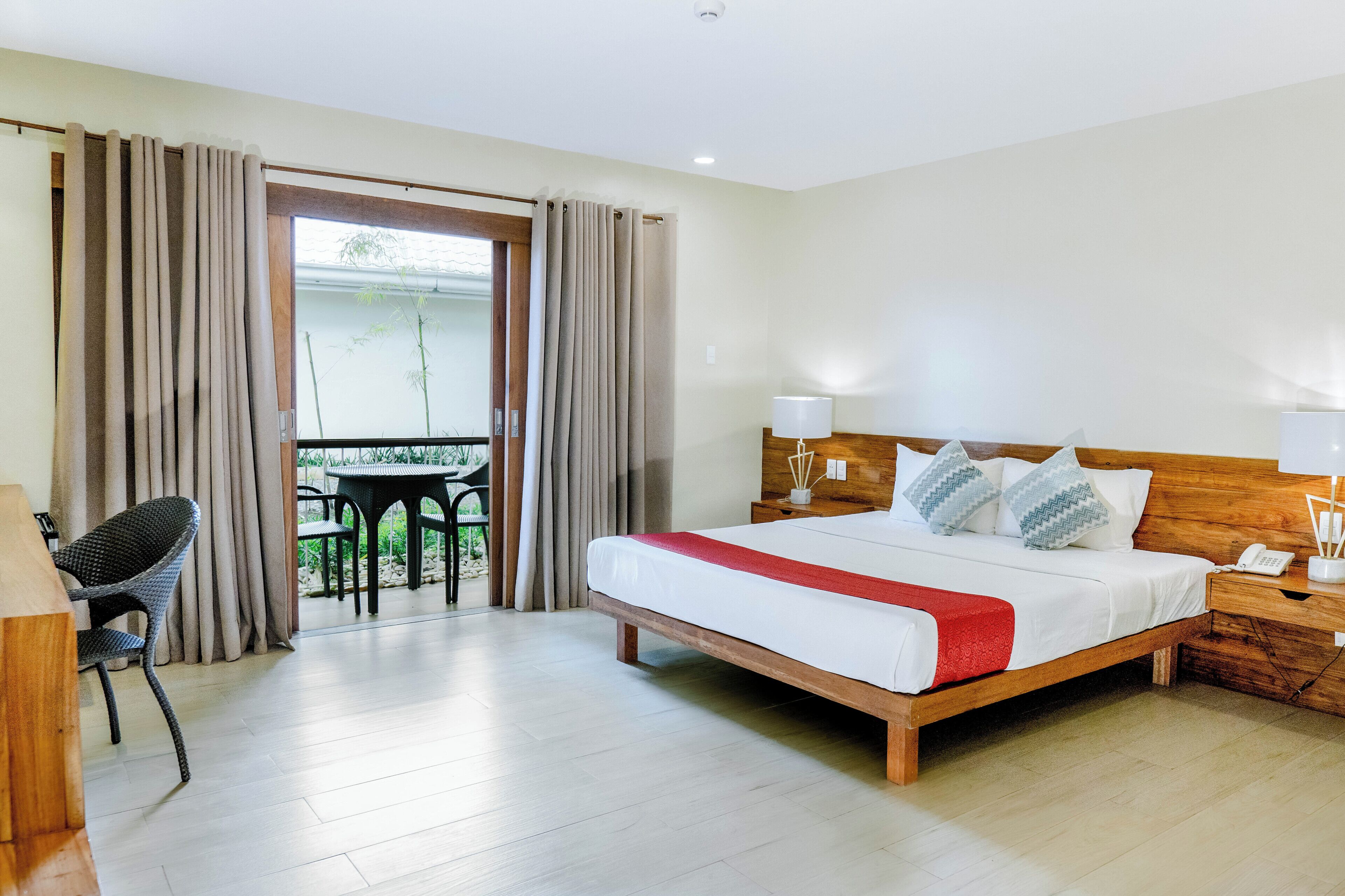 A hotel room with a bed, bedside lamps, a wooden nightstand, a chair, a table, and a view through a glass door.