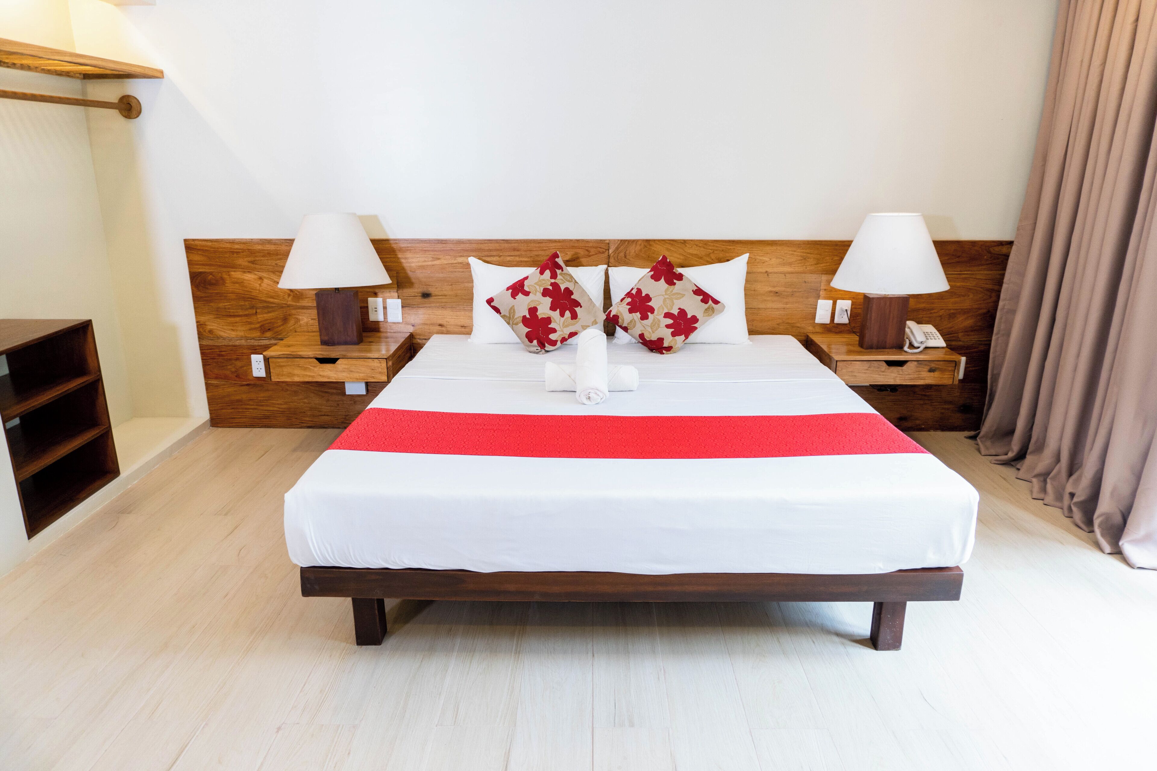Una habitación de hotel con cabecera de madera, cubrecama blanco, dos almohadas con motivos florales, lámparas de noche, teléfono y mesita de noche de madera.
