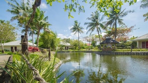 A serene pond surrounded by lush greenery and palm trees, with a thatched-roof hut and a building with a red roof in the background. - Almont Inland Resort (Butuan)