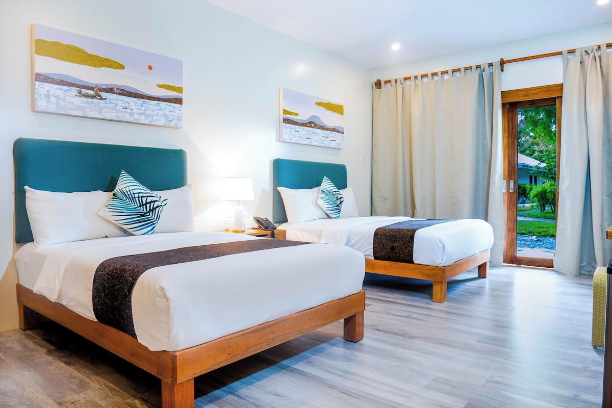 A hotel room with two beds, a wooden headboard, a large window with curtains, and artwork on the walls.