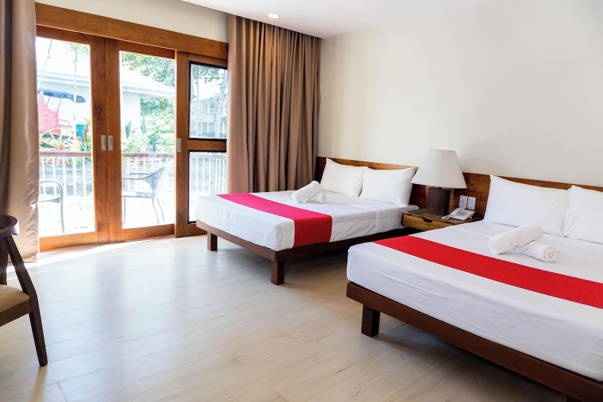 A hotel room with two beds, a wooden floor, a balcony with a view, and a nightstand with a lamp.