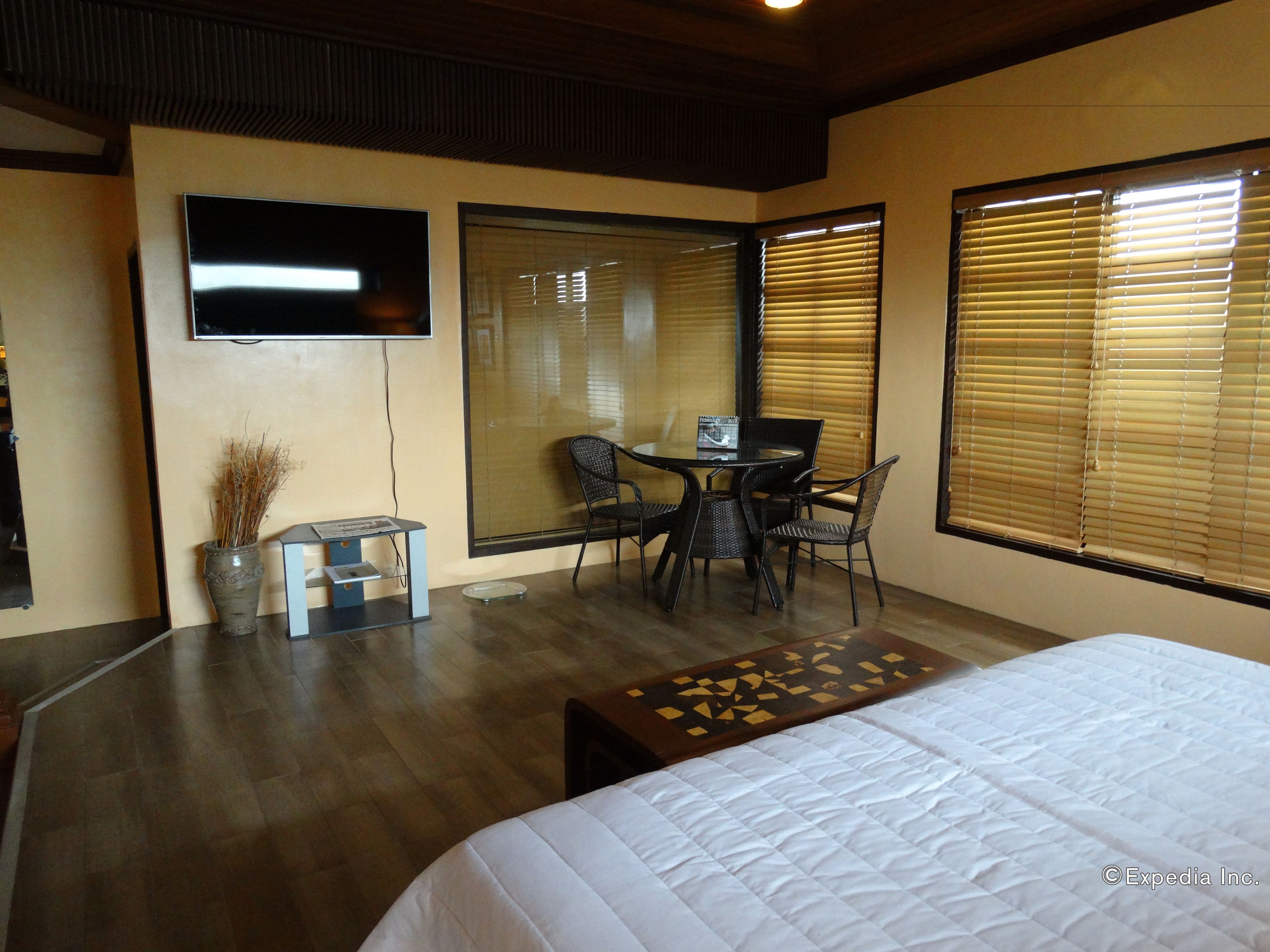 Habitación de hotel con cama, zona de comedor con mesa y sillas, televisor montado en la pared y ventanas amplias con persianas.