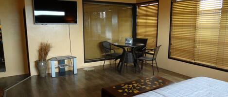 Habitación de hotel con cama, zona de comedor con mesa y sillas, televisor montado en la pared y ventanas amplias con persianas.