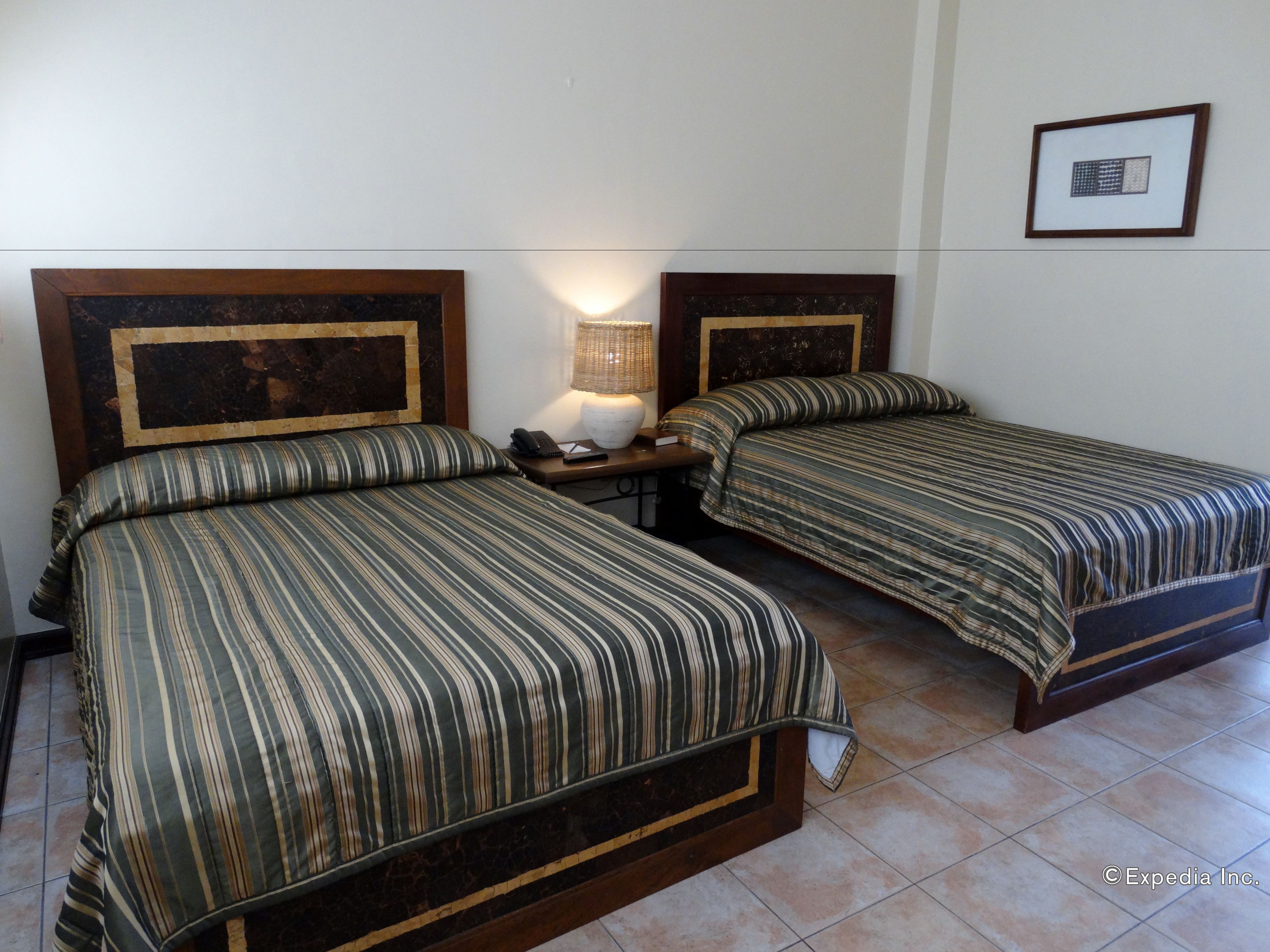 A hotel room with two beds, each with a wooden headboard and striped bedding. There is a nightstand with a lamp and a phone.
