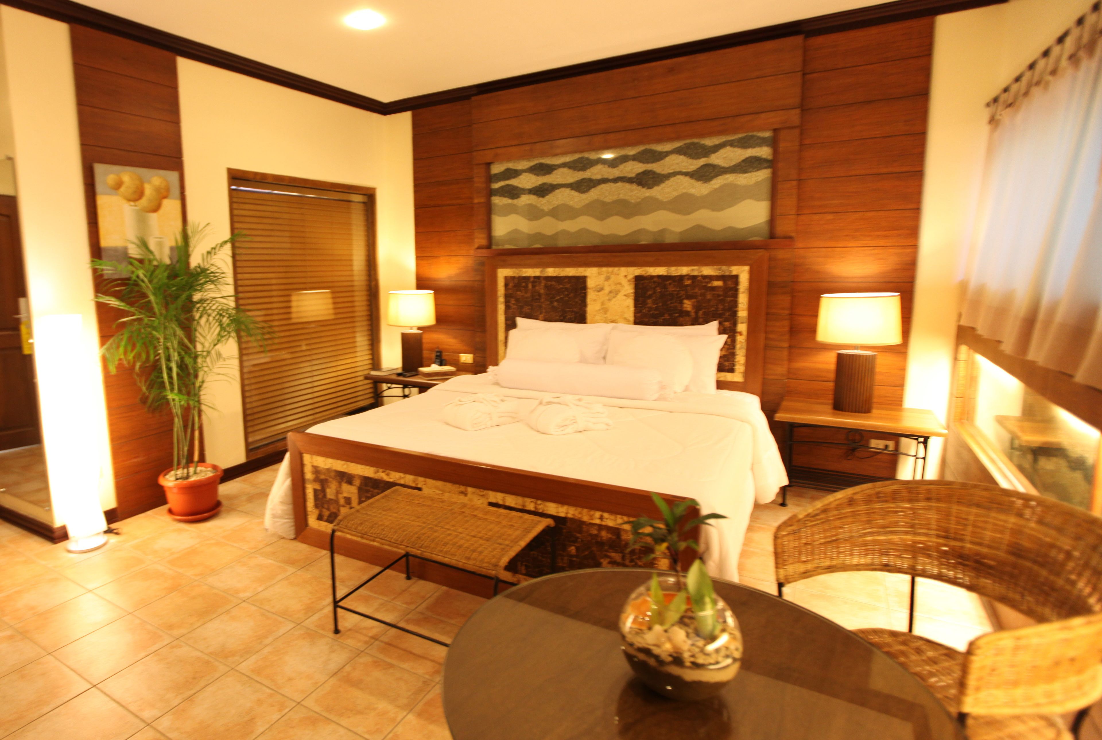 A hotel room with a large bed, two bedside tables with lamps, a potted plant, a wicker chair, and a small table with a decorative bowl.
