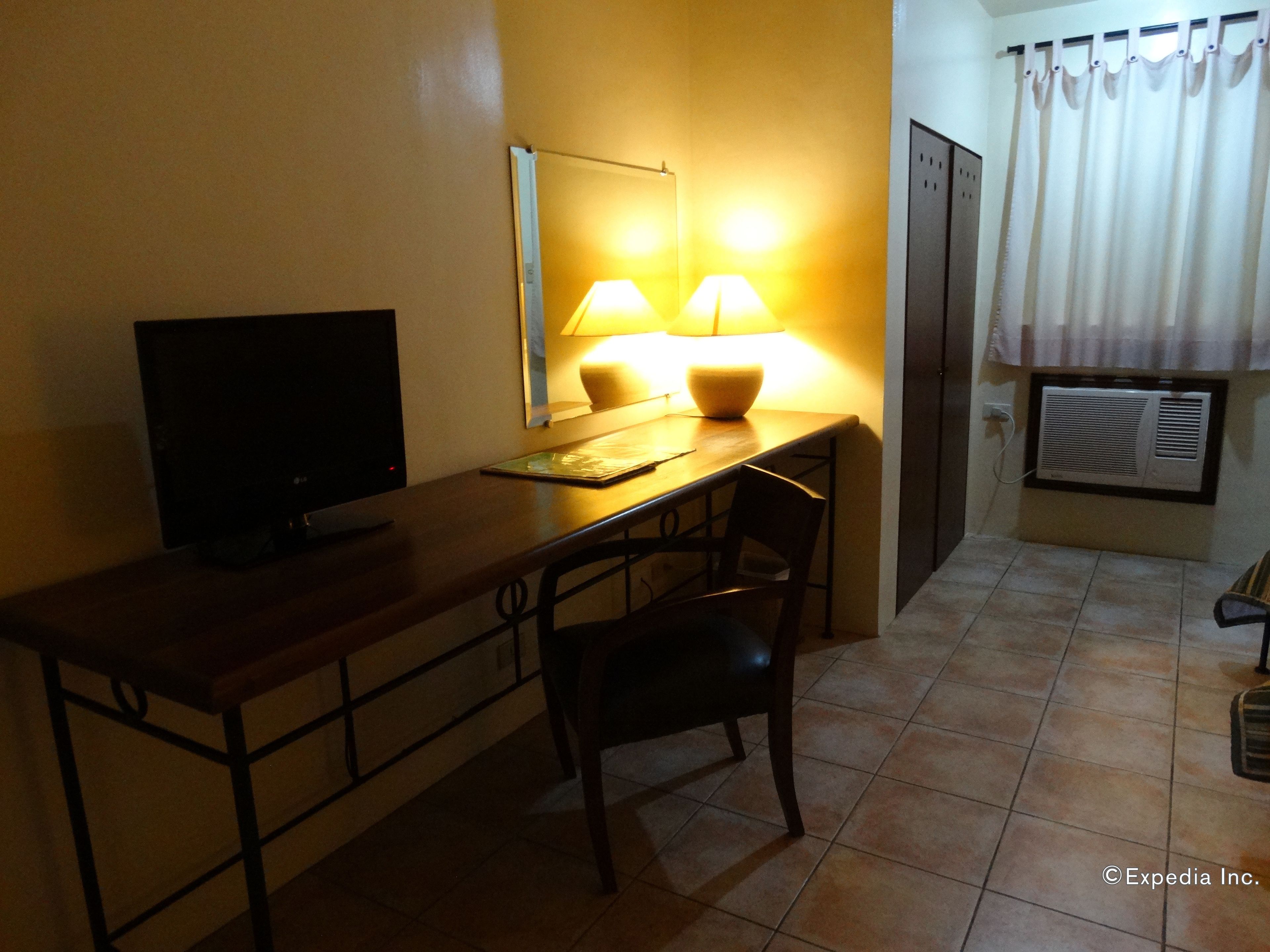 A small, dimly lit room with a desk, chair, and a television. There are two lamps on the desk, and an air conditioning unit is mounted on the wall.