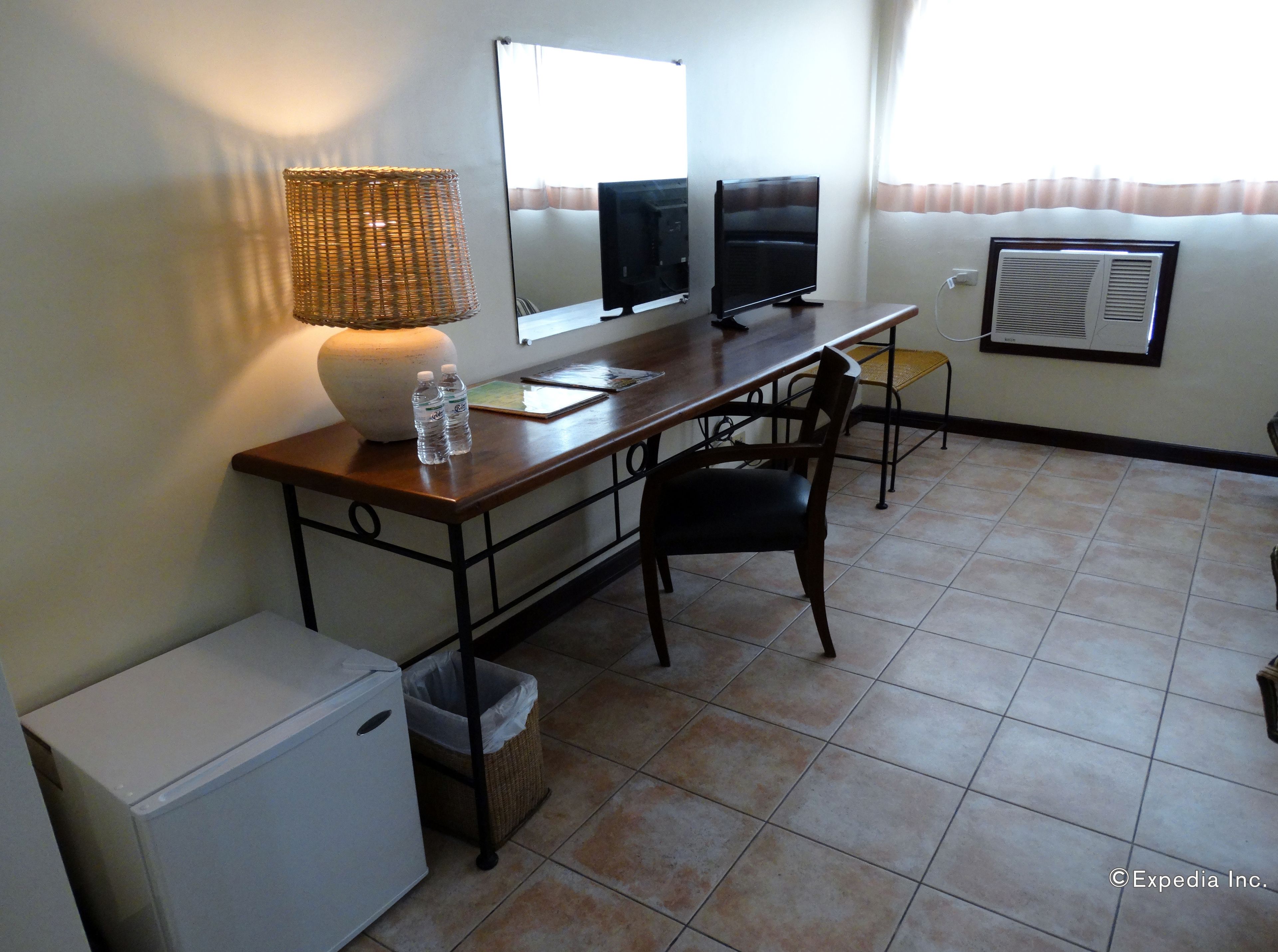 Ein Hotelzimmer mit Schreibtisch, zwei Monitoren, einer Lampe, einem Stuhl, einem kleinen Kühlschrank, einem Mülleimer und einem Fenster mit Vorhängen.