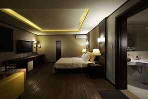 Habitación de hotel con cama, lámparas de noche, escritorio con televisor, refrigerador pequeño y baño con lavamanos y espejo.