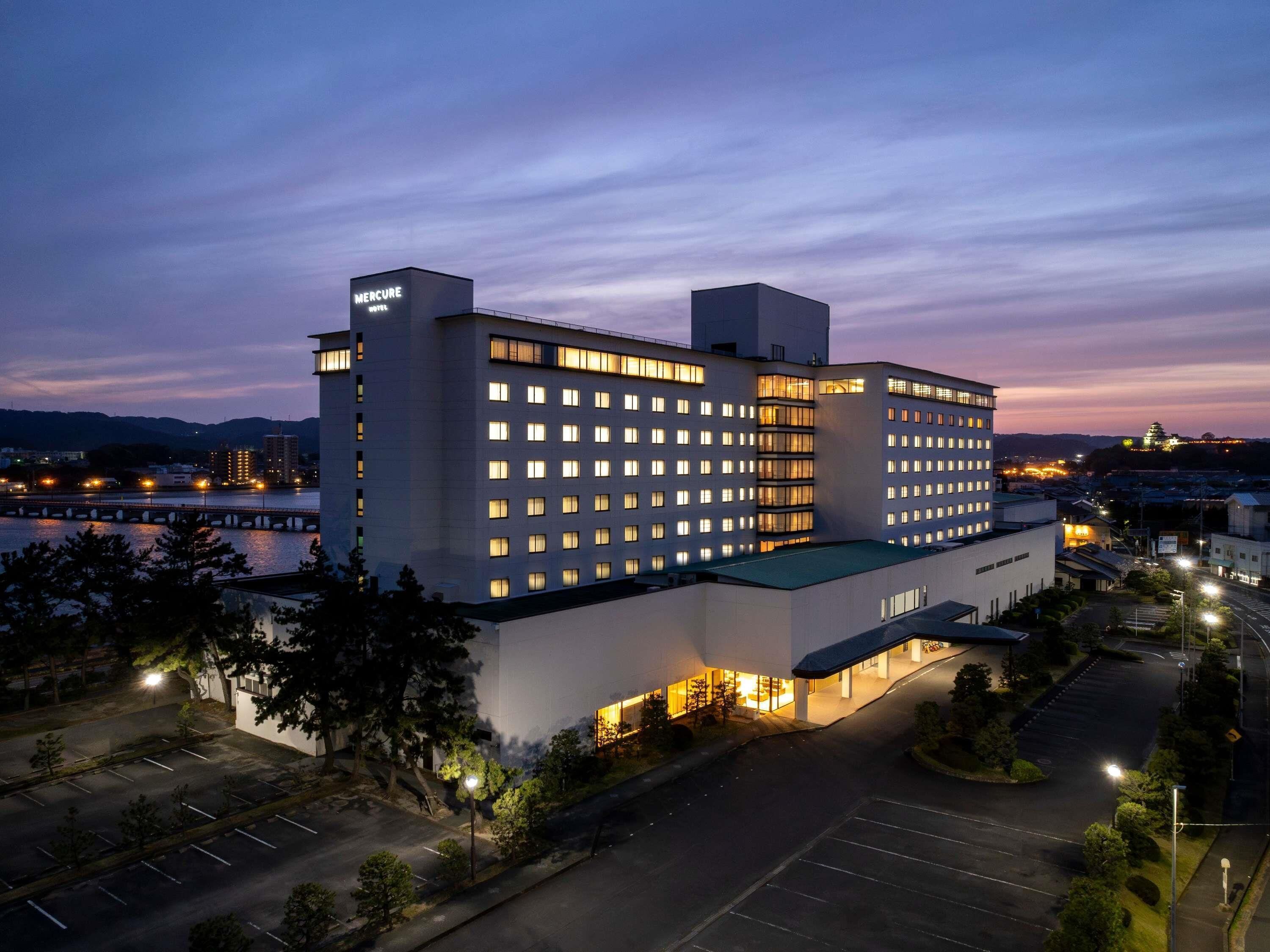 A multi-story hotel with a modern design, featuring a large parking lot and a view of a river and mountains in the background.